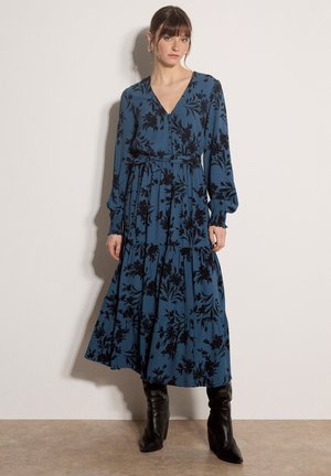 Frau trägt ein langes blaues Kleid mit schwarzem Blumenmuster und langen Ärmeln, kombiniert mit schwarzen hochhackigen Stiefeln, steht an einer schlichten Wand.