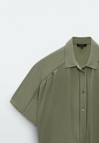 Chemise kaki à manches courtes avec boutons, col et détails cousus décoratifs le long des manches et de la patte de boutonnage.