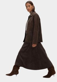 Giacca in suede marrone con colletto a punta e chiusura con bottone, abbinata a una lunga gonna marrone e stivali alti fino al ginocchio con tacco marroni.