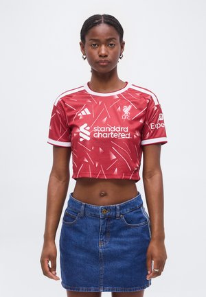 Mujer que lleva una camiseta roja corta del Liverpool FC con logotipos y una falda vaquera azul, de pie contra un fondo blanco.