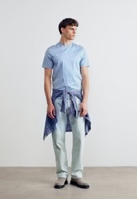 Νεαρός άνδρας όρθιος, φοράει ανοιχτό μπλε t-shirt, ανοιχτόχρωμο τζιν ξεβαμμένο, μπεζ μπότες, με δεμένο πουκάμισο καρό γύρω από τη μέση του, μπροστά σε απλό φόντο.