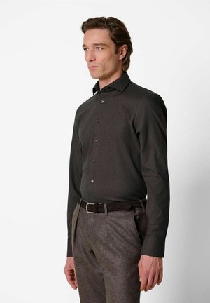 Man draagt een donker overhemd met knopen, ingestopt in grijze broek met een zwarte riem, staand tegen een effen witte achtergrond.