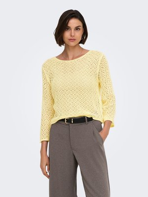 Femme portant un pull en maille texturée jaune clair à manches trois-quarts et un pantalon gris taille haute avec une ceinture noire.
