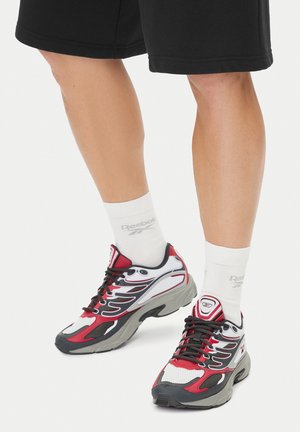 Czerwono-białe sportowe buty z czarnymi akcentami, z siateczkowego materiału, z szarą, teksturowaną podeszwą i czarnymi sznurowadłami. Noszone z białymi skarpetkami.