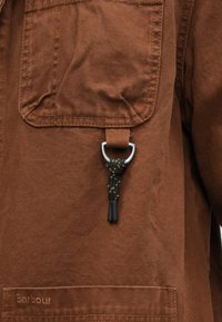 Pruun lõuendist jakk, millel on rinnatasku, musta- ja kollasevärviline nöörist aas metallist D-rõngaga ning peen logo detail ääres.