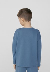 Modrá dlouhá košile s dlouhými rukávy vyrobená z měkké tkaniny s volným střihem, s kulatým výstřihem a spadlými rameny, zobrazená zezadu.