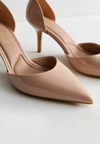 Beige gepatenteerd leren hoge hakken met spitse neuzen, een slank ontwerp en een subtiele ronding bij de hak. Glad textuur met minimale accenten.