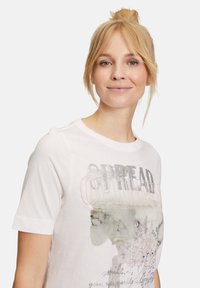 T-shirt bianco in cotone con maniche corte, scollo rotondo e un design grafico con la scritta "SPREAD OPTIMISM" in lettere argentate lucide e accenti pastello.