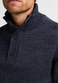 Prim-plan al unui bărbat îmbrăcat cu un pulover tricotat bleu închis, cu un guler înalt cu nervuri și două nasturi negri aproape de gât.