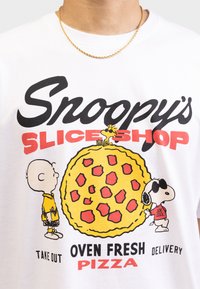Hvid t-shirt med "Snoopy's Slice Shop" grafik, der viser en gul pizza og karakterer. Teksten inkluderer "Ovnfrisk Pizza," "Take Out," og "Levering."