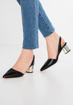 Nogi w niebieskich dżinsach i czarnych skórzanych sandałkach na obcasie typu slingback, z ostrymi noskami i złotymi blokowymi obcasami na białej podłodze.