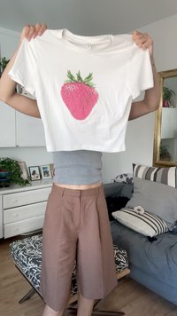 Tricou alb scurt, cu un grafic mare de căpșună roz și frunze verzi; expus pe un fundal de îmbrăcăminte casual.