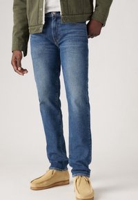 Blue denim jeans with a straight leg fit, featuring classic five-pocket design and subtle fading. Paired with beige moccasin-style shoes.