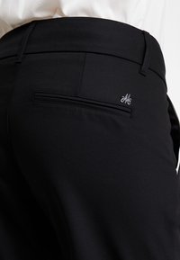 Czarne spodnie o gładkiej fakturze, z dyskretną tylną kieszenią i małym metalowym akcentem w postaci logo. Dopasowany krój o eleganckim wyglądzie.