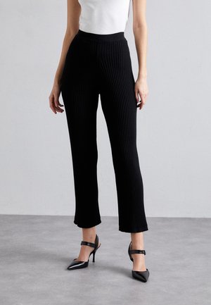 Black ribbed knit pants with a wide elastic waistband, slightly flared hem, paired with black pointed-toe heels featuring ankle straps.