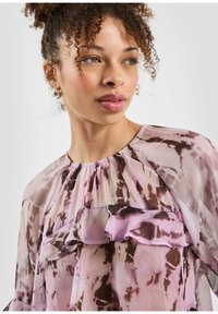 Doorzichtige roze blouse met bruine, abstracte patronen, voorzien van een gerimpelde halslijn en zachte textuur. Krullend haar en oorbellen voegen detail toe.