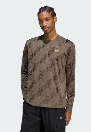 Uomo con capelli intrecciati che indossa una maglia marrone a maniche lunghe con un motivo e pantaloni neri con un logo sulla tasca.
