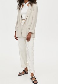 Veste beige à manches retroussées, top court blanc avec bretelles croisées, pantalons blancs retroussés, et sandales noires avec accents circulaires blancs.