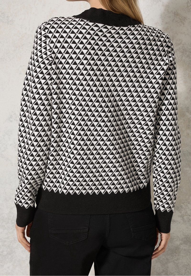 Femme portant un pull à motif géométrique noir et blanc avec des poignets et un ourlet noirs, associé à un pantalon noir, vue de dos.