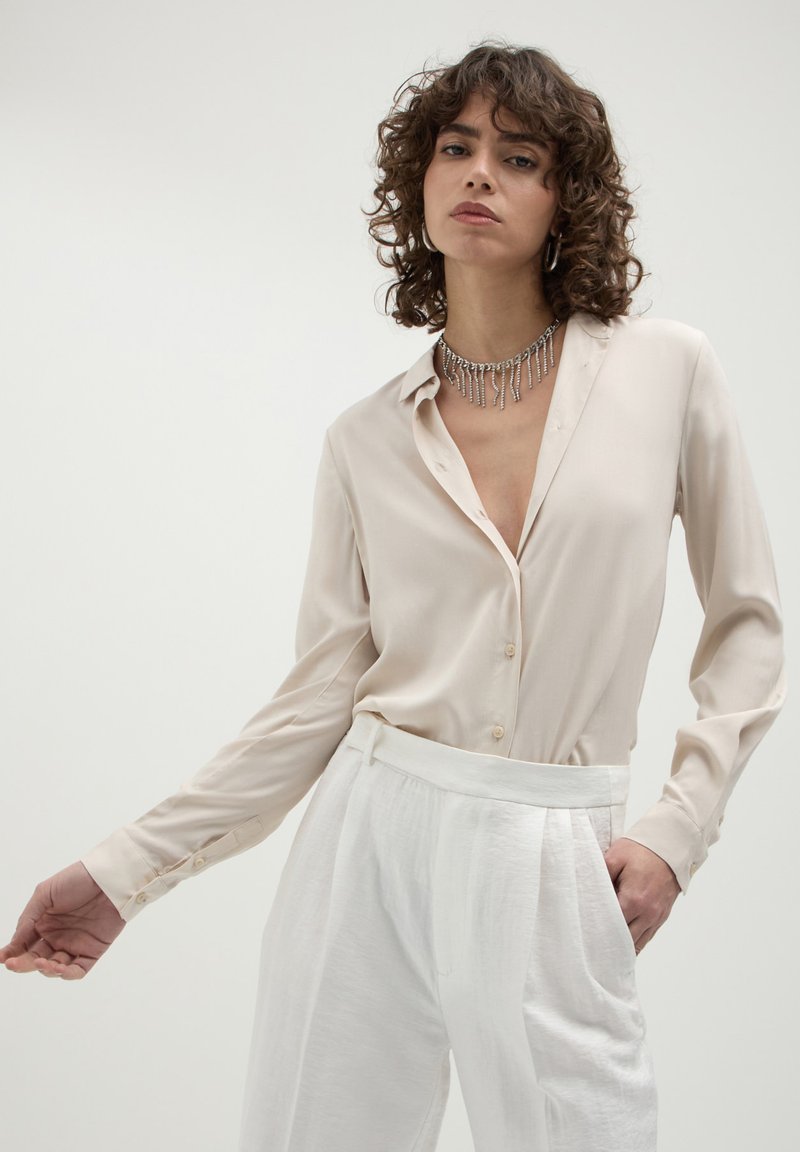 Donna con capelli ricci che indossa una blusa beige con bottoni, pantaloni bianchi a vita alta, orecchini a cerchio in argento e una collana pendente in argento.
