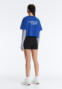 Femme aux cheveux tressés, de dos, portant un t-shirt bleu court avec un texte, manches longues rayées, short noir, chaussettes blanches et baskets.