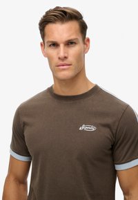 Camiseta de algodón marrón con acentos azul claro en las mangas, con un pequeño logo blanco de "Superdry" en el pecho. Corte clásico, escote redondo.