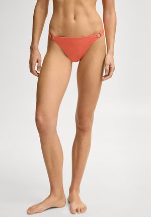 Kobieta nosząca koralowe dół bikini z metalowymi pierścieniami po bokach, stojąca boso na jednolitym, jasnym tle.