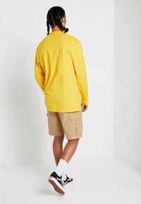 Camicia a maniche lunghe gialla in cotone, abbinata a pantaloni cargo beige. Calzini bianchi e scarpe slip-on nere completano l'outfit.