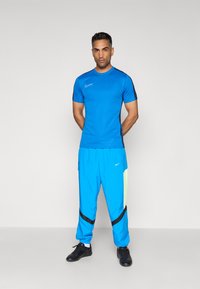 Camisa atlética azul de manga corta con acentos negros, combinada con pantalones joggers azules que tienen un panel verde neón y rayas negras. Zapatillas deportivas negras.