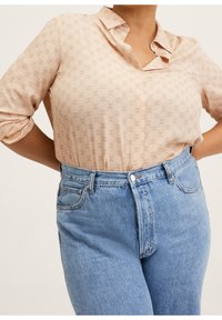 Femme portant un chemisier beige à motifs rentré dans un jean taille haute bleu clair, les mains posées sur les hanches.