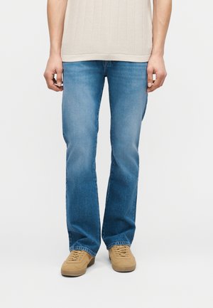 Osoba ubrana w jasnoniebieskie jeansy o prostych nogawkach, beżową koszulę o fakturze oraz brązowe buty na sznurowadła, stojąca na jednolitym tle.