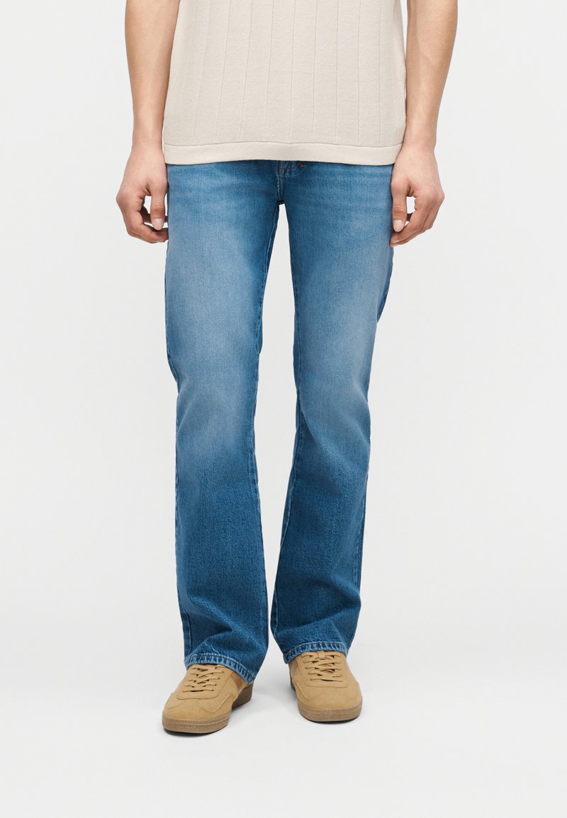 Άτομο που φοράει τζιν με ελαφρύ μπλε χρώμα και ίσια γραμμή, μπεζ υφασμάτινο πουκάμισο και ταμπά παπούτσια με κορδόνια, στέκεται μπροστά σε μονόχρωμο φόντο.