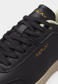 Κοντινή λήψη μαύρου δερμάτινου αθλητικού παπουτσιού με μαύρα κορδόνια, μπεζ σόλα και χρυσό λογότυπο "REPLAY" στο πλάι και στη γλώσσα.