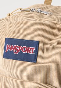 Μπεζ σακίδιο από κορδούρα με διπλό ασημένιο φερμουάρ και μπλε σκούρο ετικέτα με την επιγραφή "JANSPORT" σε κόκκινες και λευκές ραφές.