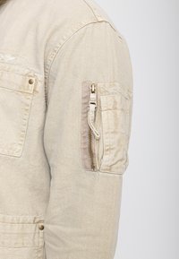 Beżowy rękaw kurtki z kieszenią na zamek błyskawiczny i kieszenią z klapką zapinaną na guzik na górnej części ramienia.