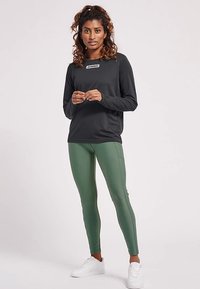 Haut noir à manches longues avec un logo blanc, associé à des leggings verts. Le modèle porte des baskets blanches. Le fond est uni.