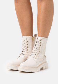 Las botas blancas hasta el tobillo cuentan con un panel tejido texturizado, punta redonda, herrajes dorados y un sistema de cordones en la parte frontal con cremallera lateral para una fácil colocación.