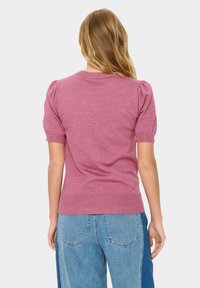 Frau mit langen blonden Haaren, die einen rosa kurzärmeligen Pullover und zweifarbige blaue Jeans trägt, steht mit dem Rücken zu einem einfarbigen Hintergrund.