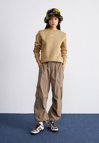 Beige Nike -collegepaita, löysät beiget taskulliset cargo-housut ja kuvioitu kalastajanhattu. Valkoiset ja mustat tennarit, joissa ruskeat korostukset. Sininen lattia.