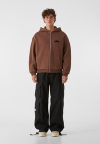 Rjav zip-up kapucar s kengursko žepom in logotipom, v kombinaciji s črnimi cargo hlačami, ki imajo več žepov in vrvice na koncu, obutimi s kremnimi športnimi copati.