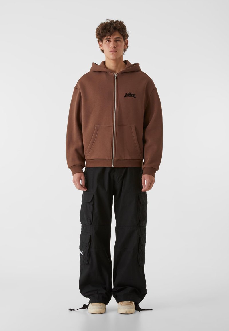 Rjav zip-up kapucar s kengursko žepom in logotipom, v kombinaciji s črnimi cargo hlačami, ki imajo več žepov in vrvice na koncu, obutimi s kremnimi športnimi copati.