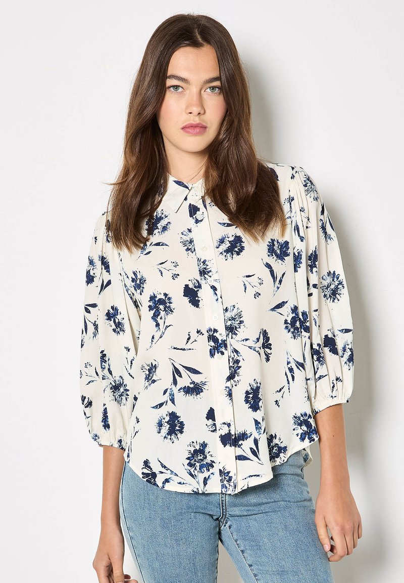 Blouse fleurie crème à motif floral bleu marine. Comprend une fermeture boutonnée à l'avant, un col et des manches bouffantes. Associée à un jean bleu clair.