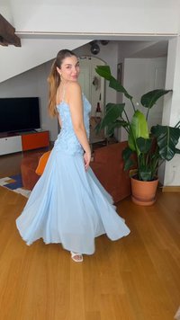 Kobieta w lekkiej, jasnoniebieskiej sukni obraca się w pomieszczeniu na drewnianej podłodze obok brązowej sofy i dużej rośliny liściastej w terakotowej doniczce.