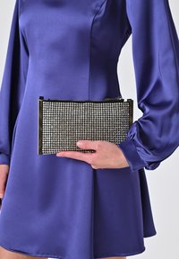 Borsa a pochette nera coperta di strass argentati, con accenti metallici e chiusura a zip, tenuta contro un vestito di raso viola.