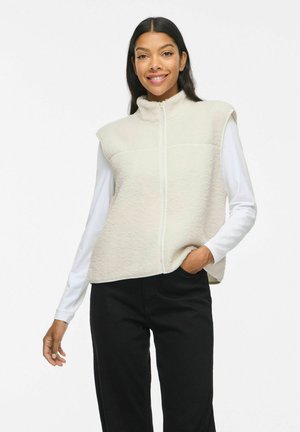 Lachende vrouw die een wit shirt met lange mouwen, een crèmekleurig mouwloos fleecevest met rits en een zwarte broek draagt, staand met één hand in de zak.