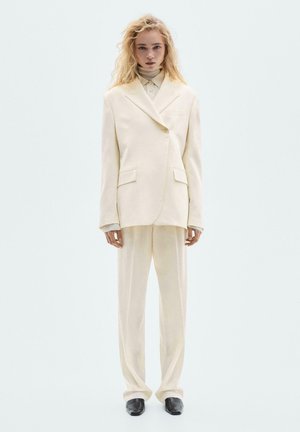 Blonde person wearing an oversized cream suit with wide-leg trousers and black square-toe shoes, standing against a light gray background.