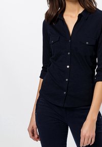 Chemise bleu marine à boutons avec deux poches poitrine, boutons-pression en argent et manches retroussées. Fabriquée en tissu doux et texturé.