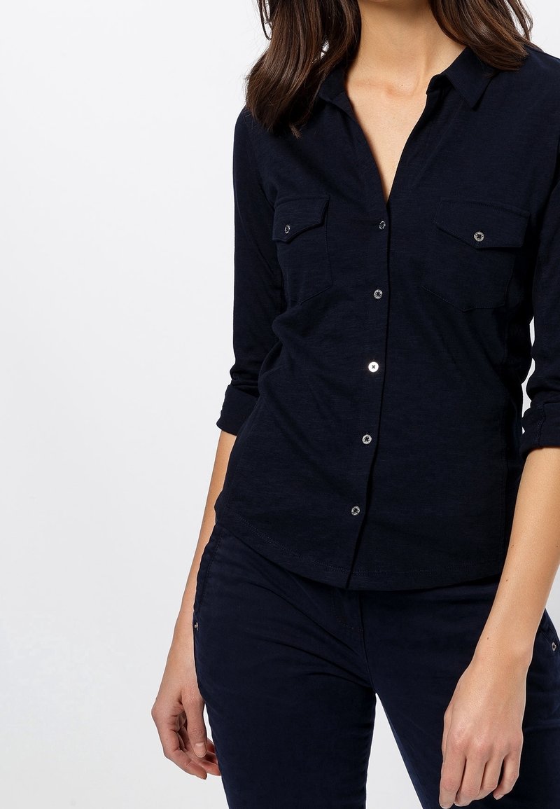 Chemise bleu marine à boutons avec deux poches poitrine, boutons-pression en argent et manches retroussées. Fabriquée en tissu doux et texturé.