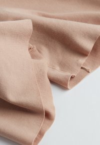 Blødt, lysebrunt stof med synlige sømme og foldede kanter på en hvid overflade, der fremhæver tekstilens tekstur og sømme.