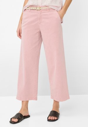 Kvinde iført lyserøde bukser med vide ben og beige bælte, kombineret med sorte flade sandaler med åbne tæer på hvid baggrund.